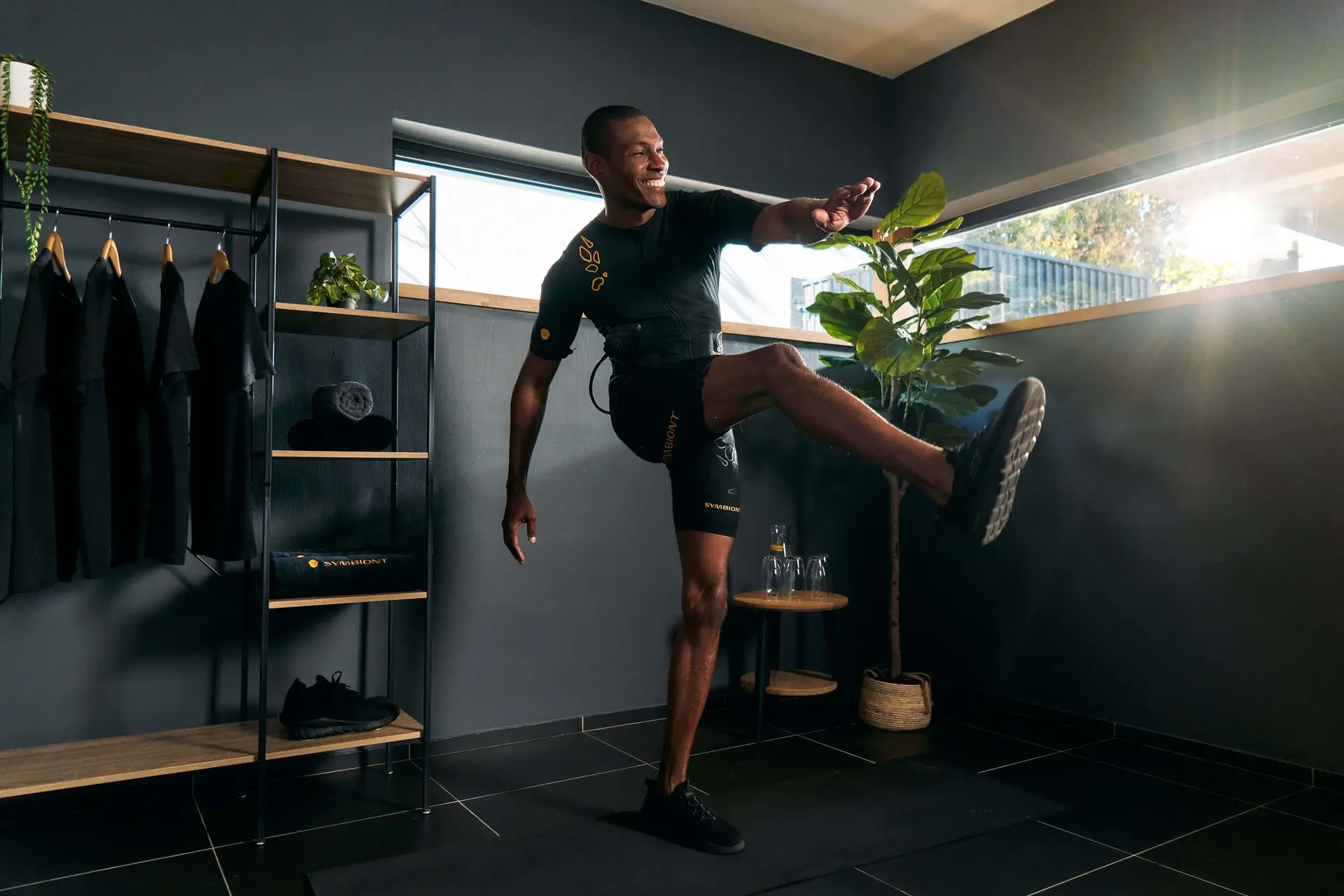 SYMBIONT EMS Experience - L'avenir du fitness avec une stimulation musculaire électrique avancée Un homme en combinaison SYMBIONT EMS effectue un exercice de levée de jambe. La pièce est baignée de lumière. Cette image symbolise l'avenir de l'entraînement EMS et du fitness.
