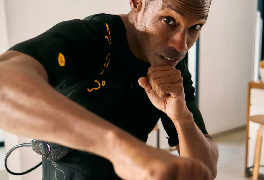 Maximisez votre entraînement avec le SYMBIONT Smart Training System, un système d'entraînement EMS haut de gamme et futuriste Une personne en combinaison EMS SYMBIONT effectue des mouvements de boxe, avec une expression faciale concentrée. Cette image représente une expérience d'entraînement avancée.