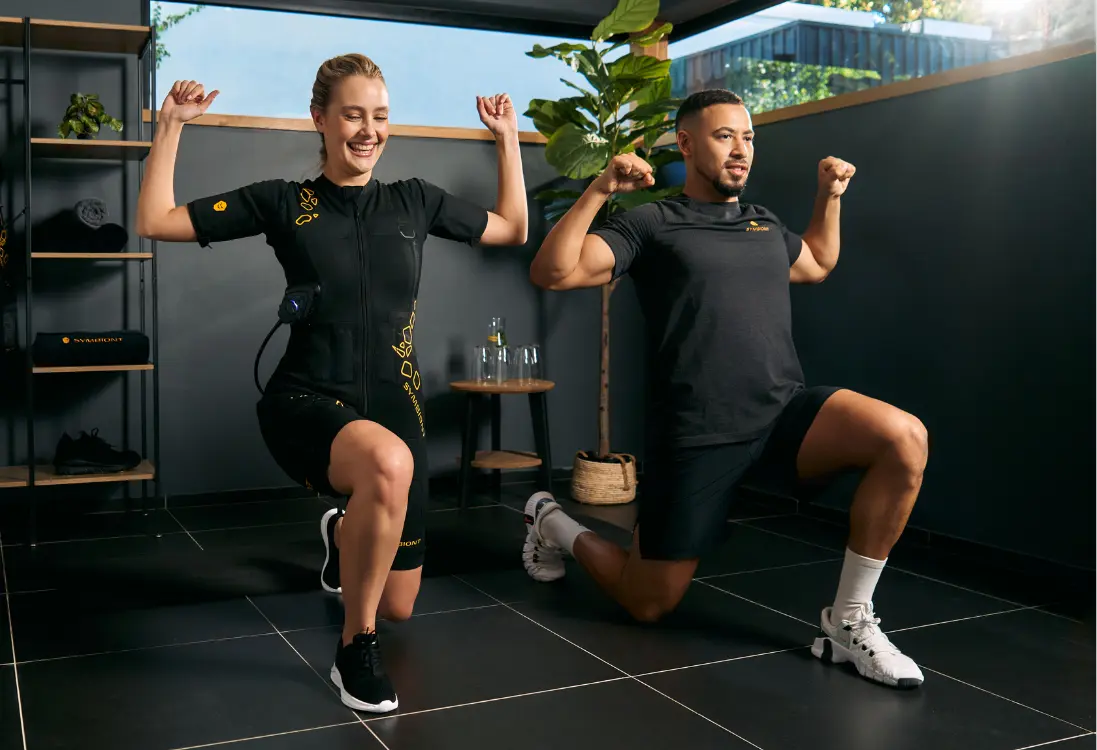 Entraînement EMS personnel ultime - S'entraîner individuellement avec SYMBIONT EMS Une femme en combinaison EMS effectue des exercices sous la direction d'un entraîneur professionnel afin de garantir un entraînement efficace et personnalisé. Elle s'entraîne dans une salle à l'aménagement minimaliste et baignée de lumière.